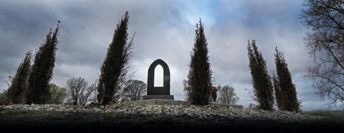Gray winter skies loom over the prominence of a monument. It's surrounded by tall, thin evergreens. The ground is covered with hand-sized stones.