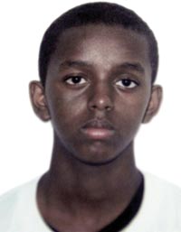 An official-looking portrait of a boy, looking perhaps old enough to be in high school. His hair is close-cropped and his ears stick out. He wears a white T-shirt. He does not smile.