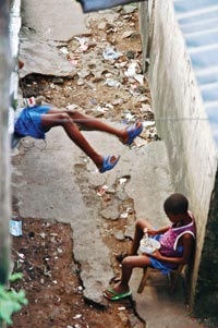 Two children sit in a trash-strewn alley. Both wear shirts and plastic sandals. One is eating out of a white, plastic bowl.