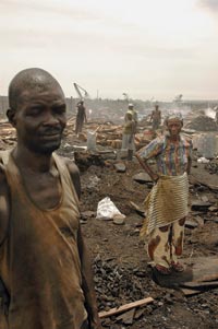 Bedraggled figures dot a smoky landscape. In the foreground stands a man, eyes closed, wearing a soot-stained steeleveless t-shirt. Behind him stands a woman, hands on her hips, wearing sandals and drab-colored clothes. A small pile of chunks of wood is on the ground between them. Smoke rises here and there.