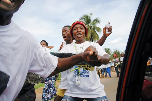 A sparse crowd, marching down the street, addresses their chants to the photographer, who is inside of a car. The woman at the center of the frame has her mouth open, speaking. She wears a “George M. Weah for PRESIDENT” T-shirt and jeans.