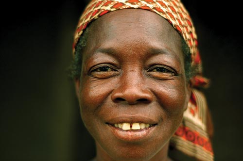A portrait of a middle-aged woman. She looks directly at the camera, smiling. There is a gap between her front teeth. Below her headscarf can be seen a tangle of graying black hair. The skin around her eyes crinkles up.