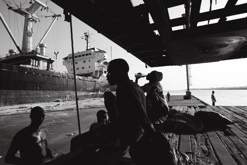 Several men rest in a patch of shade on a pier. Behind them looms a large factory trawler.