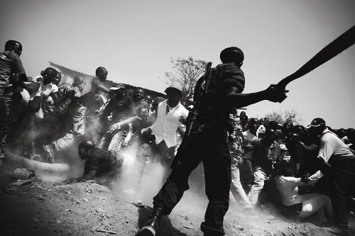A menacing-looking soldier swings a machete to clear a crowd. Dozens of people have jumped back or dived for the ground to escape the blade, raising a cloud of dust.