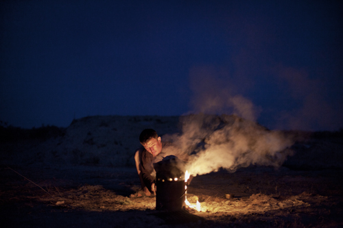 Nyrbek cooks dinner for himself and his brother Islam over an outdoor fire pit at night on a deserted piece of land.