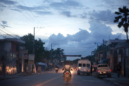 A city street at dusk with motorcycles on the roadway and several vehicles parked off the the right hand side.