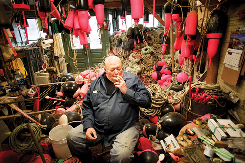 Steve Robbins in his shop, smoking a cigarette, surrounded by ropes and buoys.