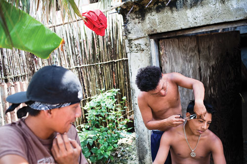 A teenage Kuna boy cuts anothers hair outside a building while a third looks on.