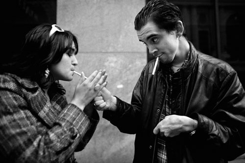 Nick, on the street, in his leather jacket, lighting a cigarette for a girl with dark hair.