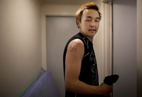 Dines standing outside the door to his apartment.  He is wearing a sleeveless shirt and has a swastika carved into his upper arm.