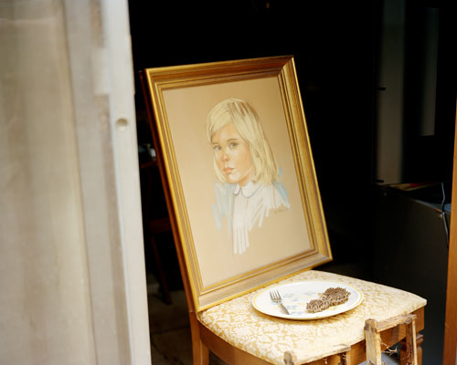 A chair with a painting of the bust of a young girl resting atop it.  On the chair is also a plate of food.