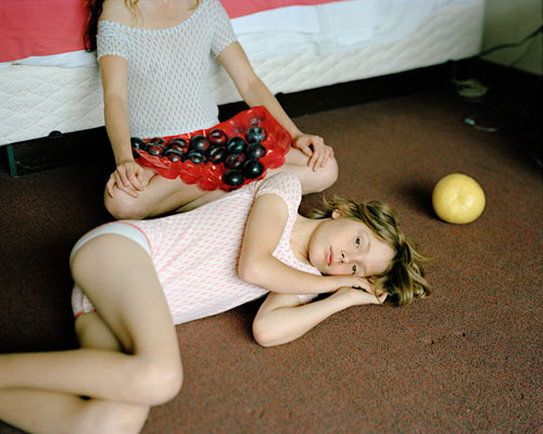 Two young girls in leotards resting on a bedroom floor.  One had several black plums in her lap.