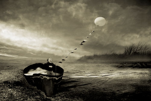 A small child stands in the water-filled hull of an old boat on a stormy day, flying a kite-like balloon on a string.
