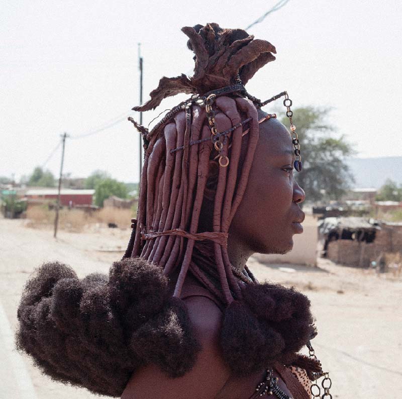 A Himba woman wears ekori, a married woman’s headpiece fashioned from cowhide, but the puffed ends of her Remy hair extensions are from China and a plastic cowrie shell dangles like a third eye.