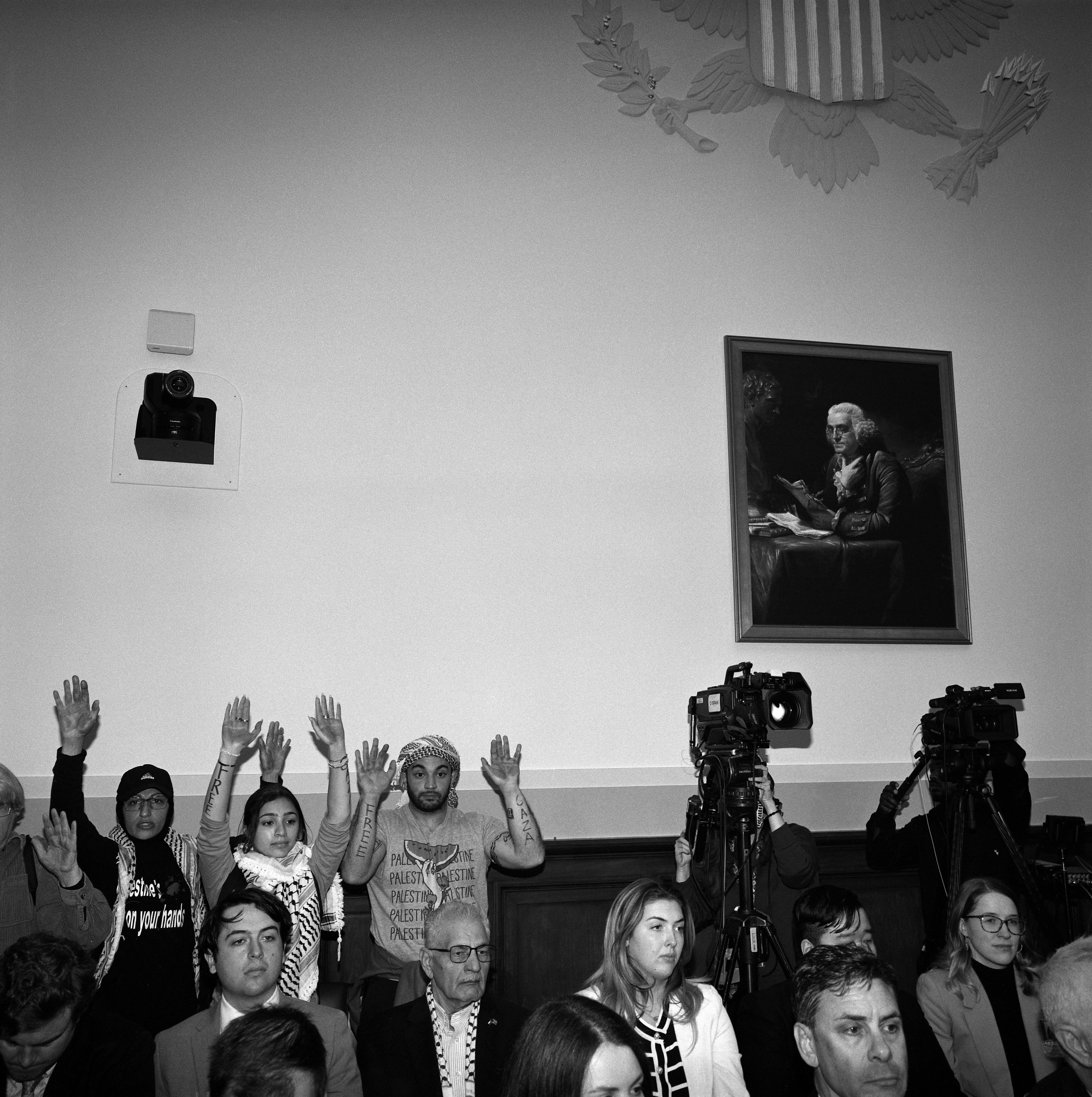 Silent protestors in back of Capitol Hill hearing with hands up in the air next to a video camera.