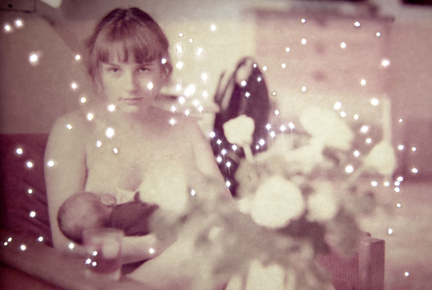 Photograph by Amy Friend of a baby being cradled by its mother. Photograph overlaid with small bright flash effects like stars or light on water.