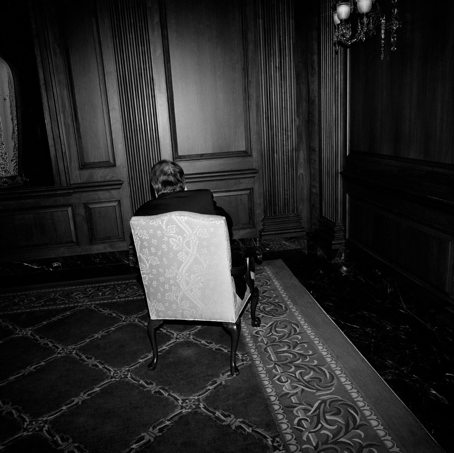 Man sits on chair in corner of room with back facing camera. 