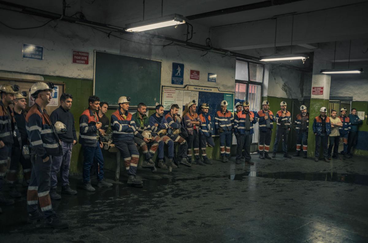 About 20 men in uniform line the perimeter of a room, appear to be listening to someone out of frame