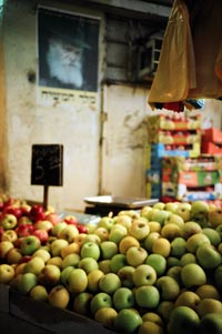 A stand is piled high with apples, plastic grocery bags hang overhead. Behind is a wall, high on which is a poster of a glowering, bearded face.