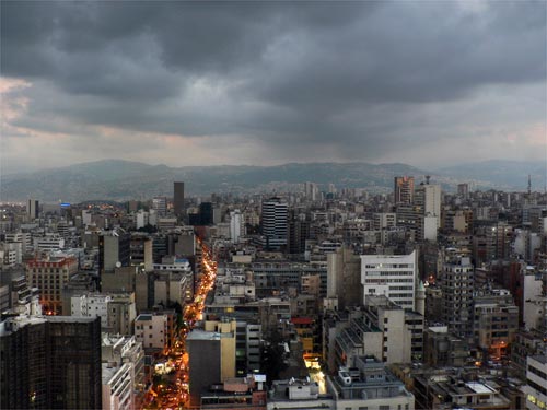 A large city at dusk, with towering buildings stretching into the distance, with a backdrop of mountains. The sky is gray, textured with heavy clouds.