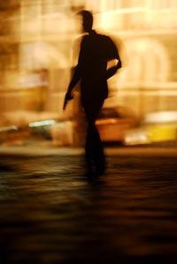 Seen in a blurred silhouette, a man walks down a street, holding a handgun at his side. It is daytime, and he is backlit.