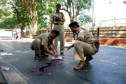 Three khakied police officers attend to a crime scene, in the form of a pool of blood in a street. Some of it has run downhill in a rivulet.