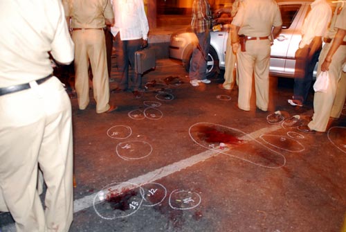 Nine people stand in and around a crime scene on an urban street. A series of numbers chalk circles on the asphalt mark apparent evidence, including bloodstains and a pair of shoes. It is nighttime.
