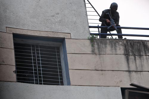 A soldier, clad all in black with a helmet and a face covering, leans over a railing on a balcony, looking down while clutching a Heckler & Koch MP5 assault weapon.