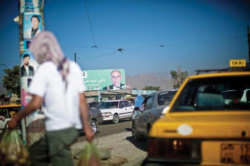 A road, crowded with cars, runs past a billboard promoting a candidate for office. A telephone pole is also plastered with posters for candidates.