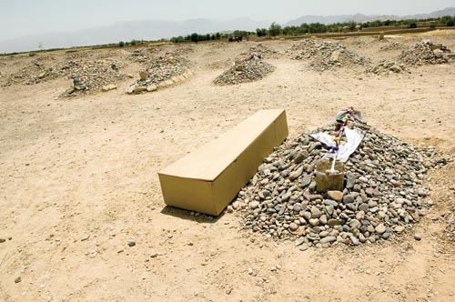 A simple coffin lies next to a grave. The grave is one of dozens. Each one consists of a pile of stones, with one large, upright headstone.