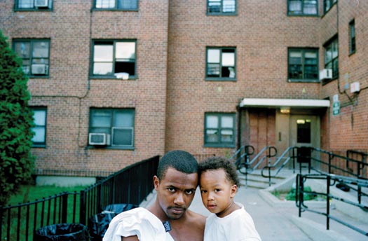 A bare-chested man, apparently in his twenties, holds a toddler in his arms. They gaze directly into the camera. Behind them is a brick housing complex.