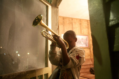 A young boy stands in the doorway to a house, playing trumpet, its bell pointed triumphantly towards the sky. He smiles slyly and looks straight at the camera.