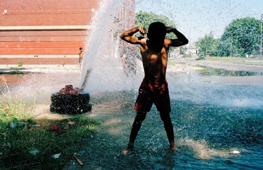 Standing before a fountaining hydrant, a boy stands ankle-deep in water, flexing his biceps in his bathing suit.