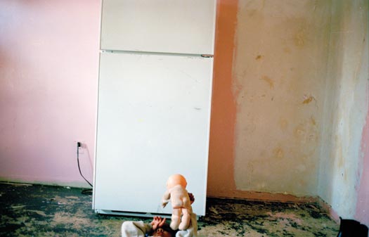 A bare room, old pink and white paint splotched on the walls, green paint peeling up from the floors. The only object within it is a lone refrigerator. A young child lies on her back on the floor, playing with a naked doll.