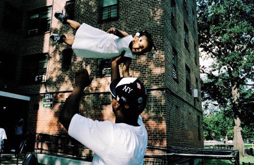 A man wearing a Yankees cap tosses a toddler into the air as she laughs. The toddler is wearinga white dress and tiny sneakers. Behind them is a large, brick public housing complex.