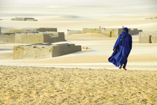 Surrounded by desert sands, a person walks towards the camera, wearing flowing blue robes and a headscarf. In the background are low, sand-colored buildings, partially buried in sand.
