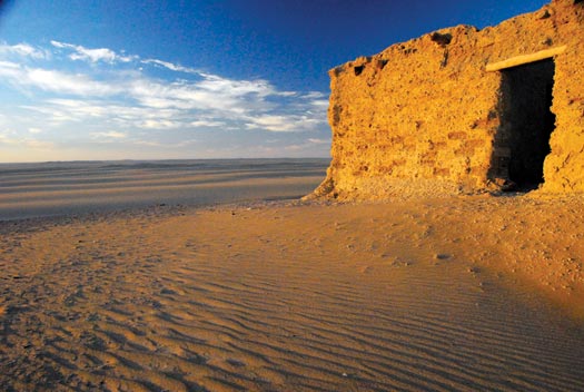A sandstone block building is surrounded by rippling desert as far as the eye can see. It has an orange cast in the long light. Overhead, the sky is blue.