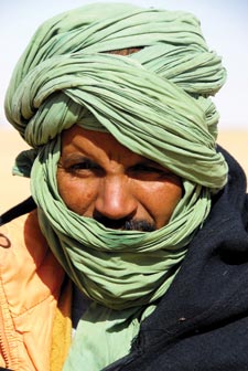A moustached man has much of his head wrapped in a green scarf. He wears a western-style woolen coat with neon orange lining and a zipper.