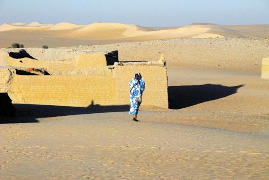 A woman wrapped in blue-and-white fabric walks across the sand, away from a low sandstone building.