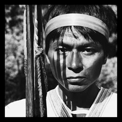 A South American man stares into the camera, his expression impassive. A narrow band of fabric encircles his head. He holds some sort of a staff, perhaps a bow, at his side.