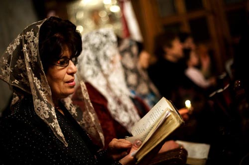 A woman reads from a small book while seated in a pew. She has a scarf covering her head.
