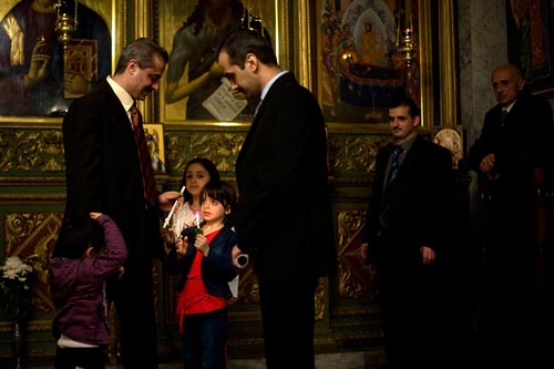 Men in suits stand as small children look at a lighted candle that one of the children holds.