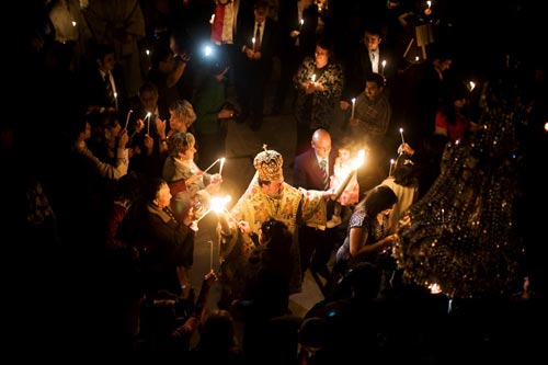 Viewed from above, a crown-wearing priest stretches his arms out, holding two candles high. Paritioners surround him, holding tapers, which they light off of his candles.