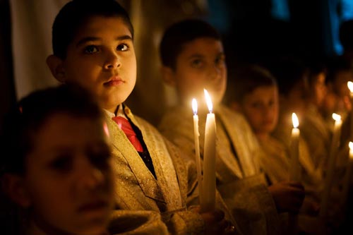A line of children, wearing robes over suits, stand holding candles that light their faces.