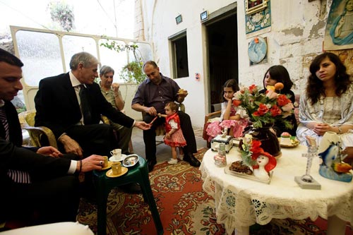 A rough-looking but well-decorated high-ceilinged room has a dozen olived-skinned family members sitting around, in various levels of finery. Some hold small cups of coffee. A toddler in a dress stands next to one man.