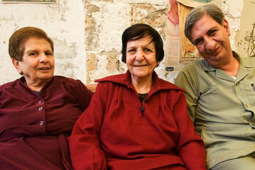 Three smiling older people look at the camera, as if for a portrait: two women and a man. Behind them, white paint is flaking off the wall.