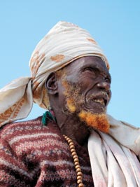 The sky a pale blue behind him, an old man squints into the sun. He has a hennaed, patchy beard, wears a scarf on his head, appears to be wearing a patterned sweater, and a long, beaded necklace is emerging from his sweater.