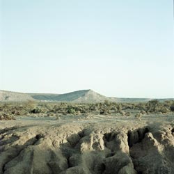 A rough expanse of desert extends into the distance. Far off is a small prominence, the plain leading to it covered in low scrub. In the foreground is deeply-furrowed land, eroded as if by a great rainstorm, though the dryness of the earth indicates it was no time recently.