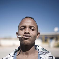 A teenaged boy, head cocked back and a smile on his lips, looks at the camera through sun-squinted eyes. A cigarette juts out of the corner of his mouth.
