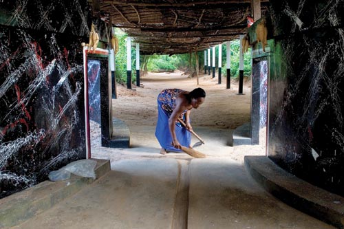 A woman wearing a blue dress stoops over while sweeping a dirt floor. She's surrounded by the cement walls and thatched roof of a shrine.